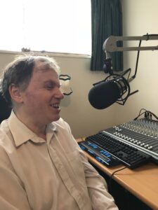 Jonathan Mosen talking into a microphone with a Braille display and studio mixer in front of him.